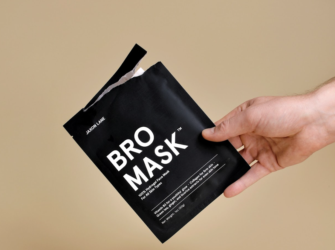 Hand holding a black mask package labeled 'Bro Mask' against a beige background