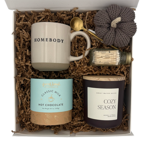Gift set with 'HOMEBODY' mug, hot chocolate mix, candle, and pumpkin in a box.