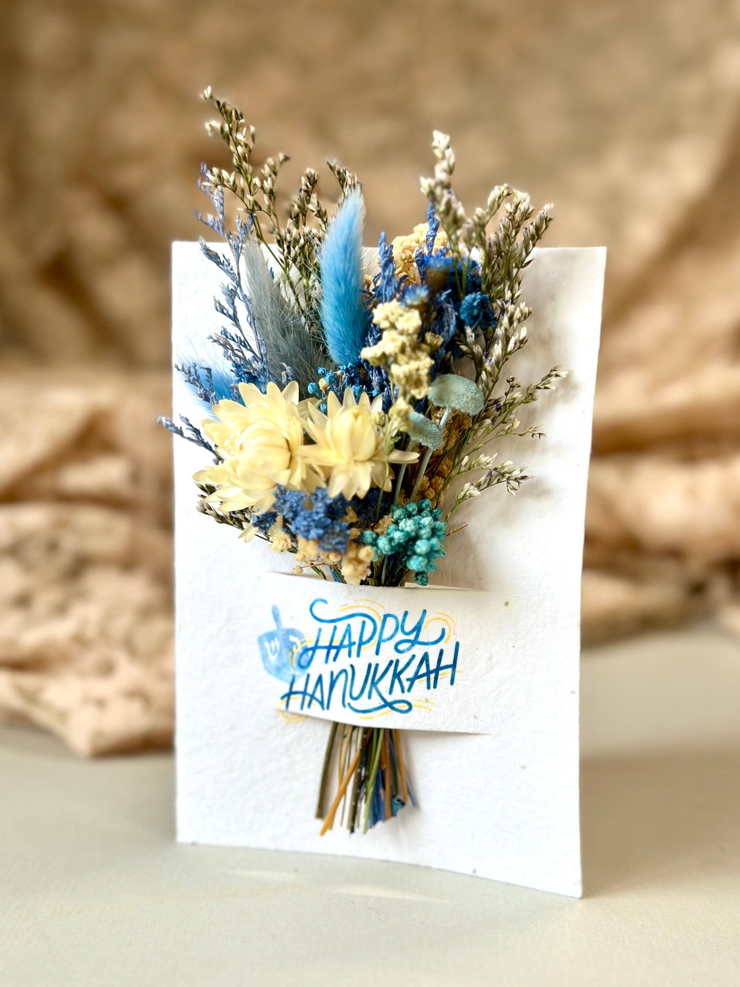 Bouquet of dried flowers on a card with 'Happy Hanukkah' text, against a beige background.