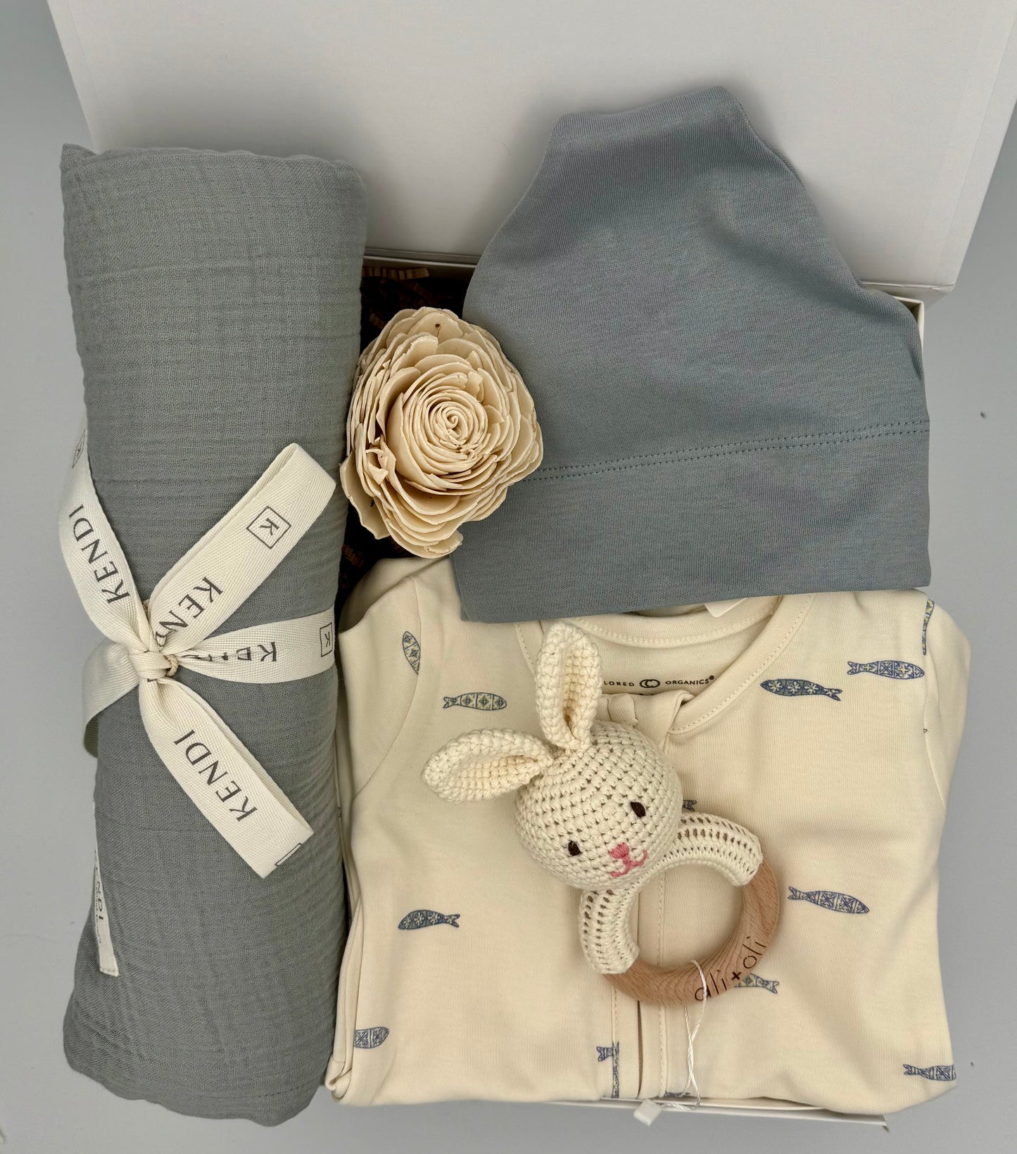 Baby clothing and用品 including a gray swaddle, blue hat, and beige outfit with a toy on a light gray background.