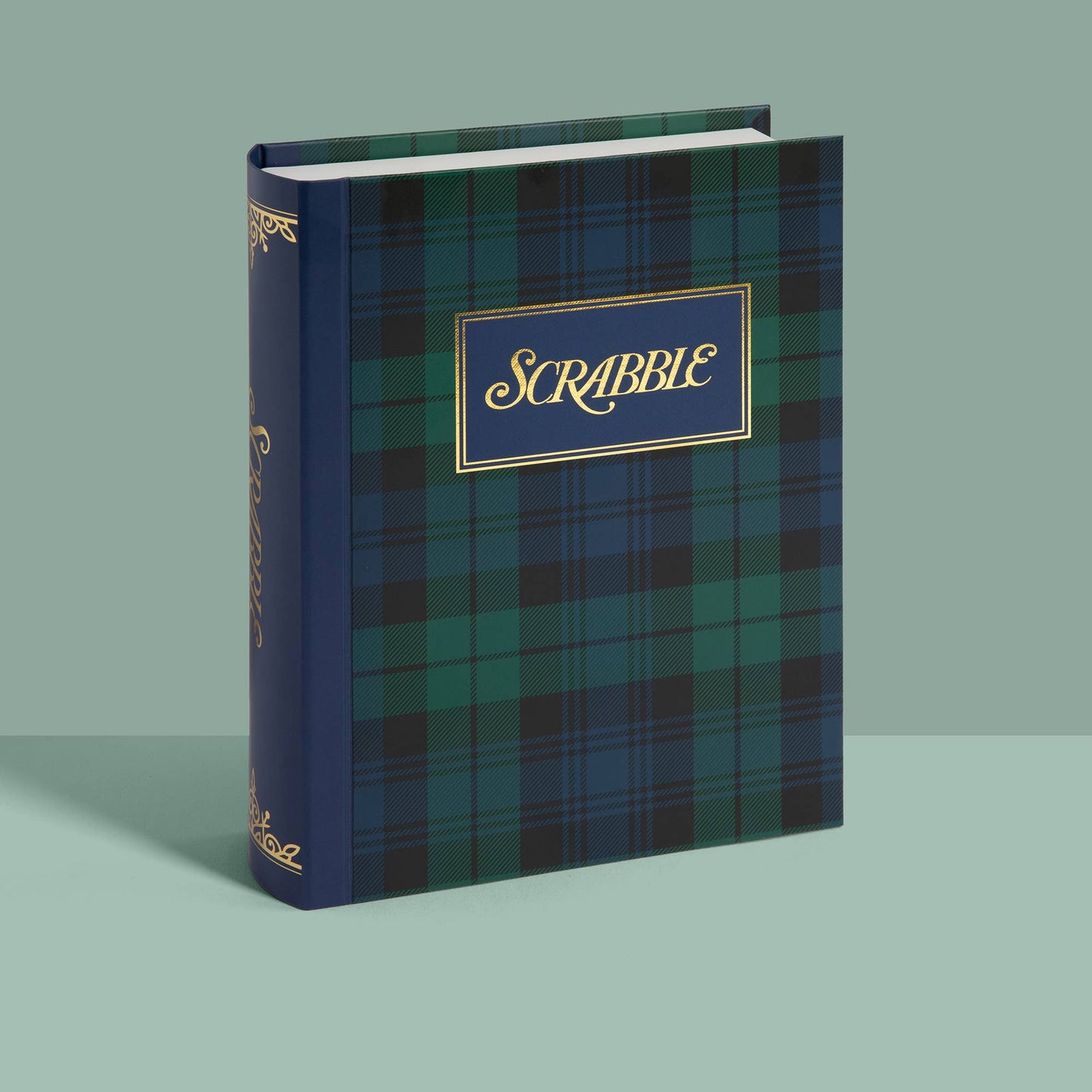 A Scrabble game box with a dark blue and green plaid cover and a navy blue spine, standing upright on a light mint-green background.