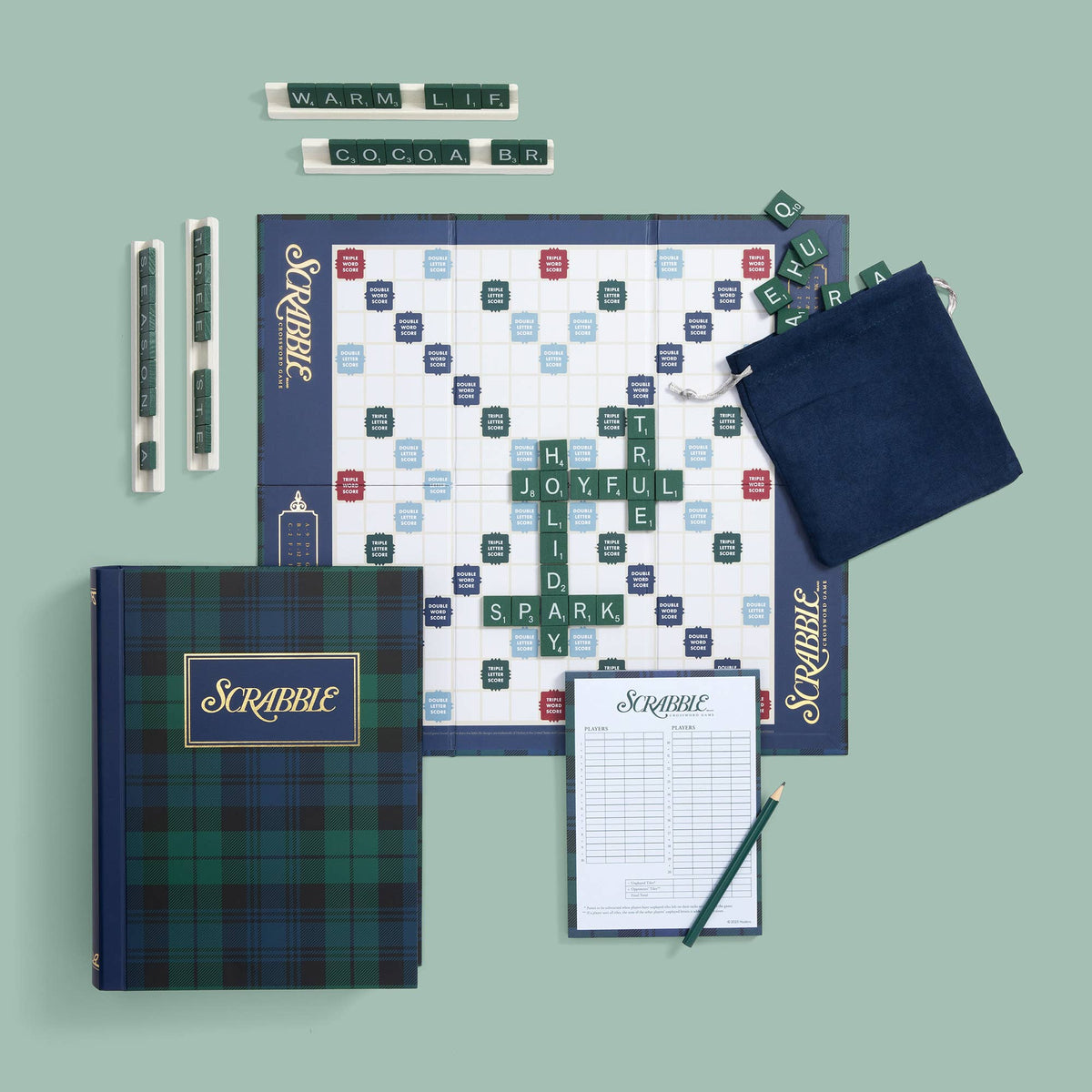 A flat-lay of a Scrabble game set on a muted sage green background, including a plaid game box, game board, word tiles, two pens, a score pad, and a navy draw bag.