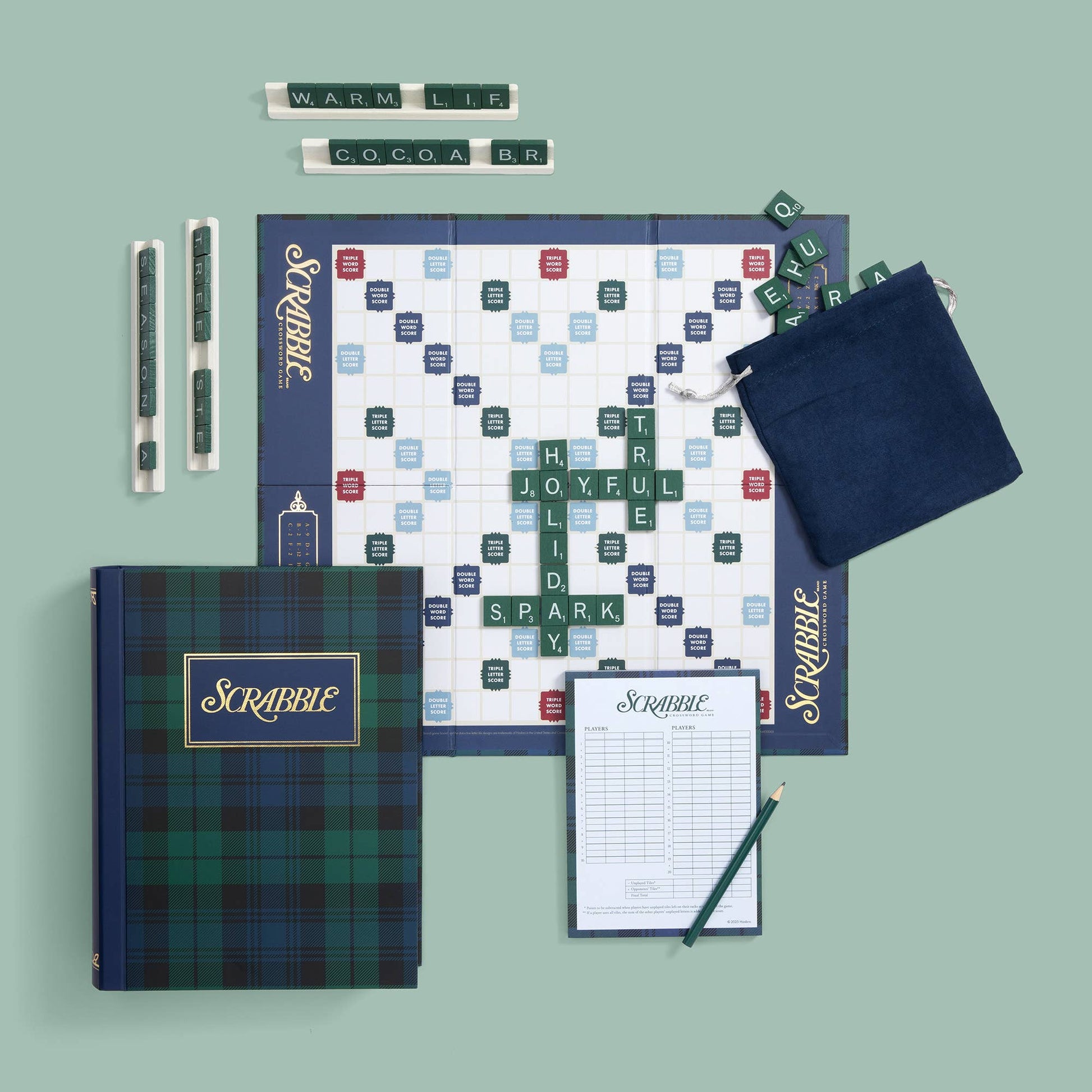 A flat-lay of a Scrabble game set on a muted sage green background, including a plaid game box, game board, word tiles, two pens, a score pad, and a navy draw bag.