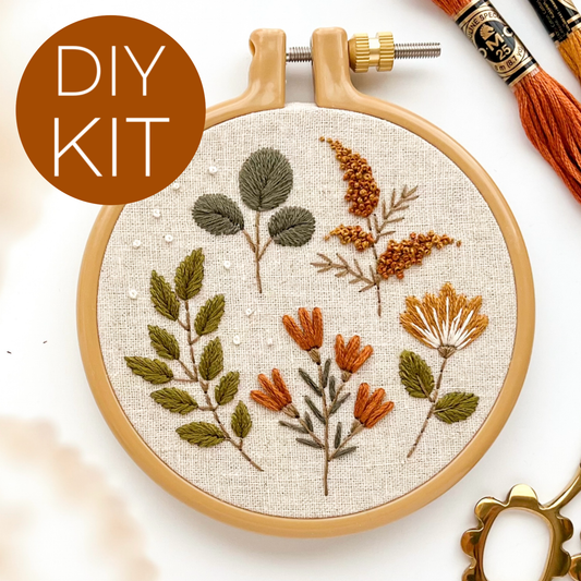 A DIY embroidery kit shown in a wooden hoop with floral stitching on cream fabric, plus thread spools and a decorative gold ring on a white background. A brown circle in the top left reads “DIY KIT.