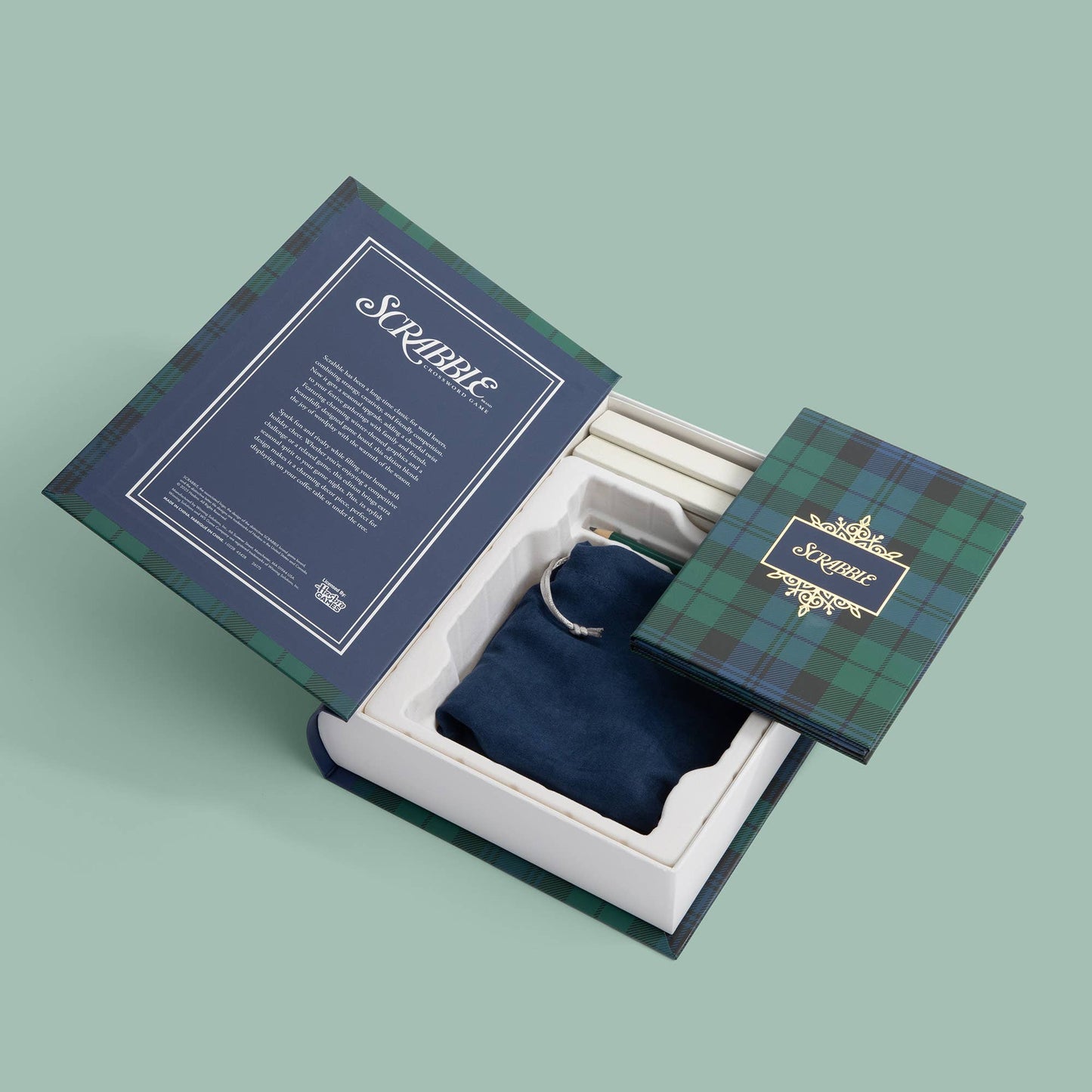 An open luxury gift box with a green tartan plaid lid, white interior, and a dark blue velvet pouch inside. The inside lid has a navy panel with white text reading 'Scruble', and the outside lid has a matching green tartan pattern with a gold 'Scruble' crest.
