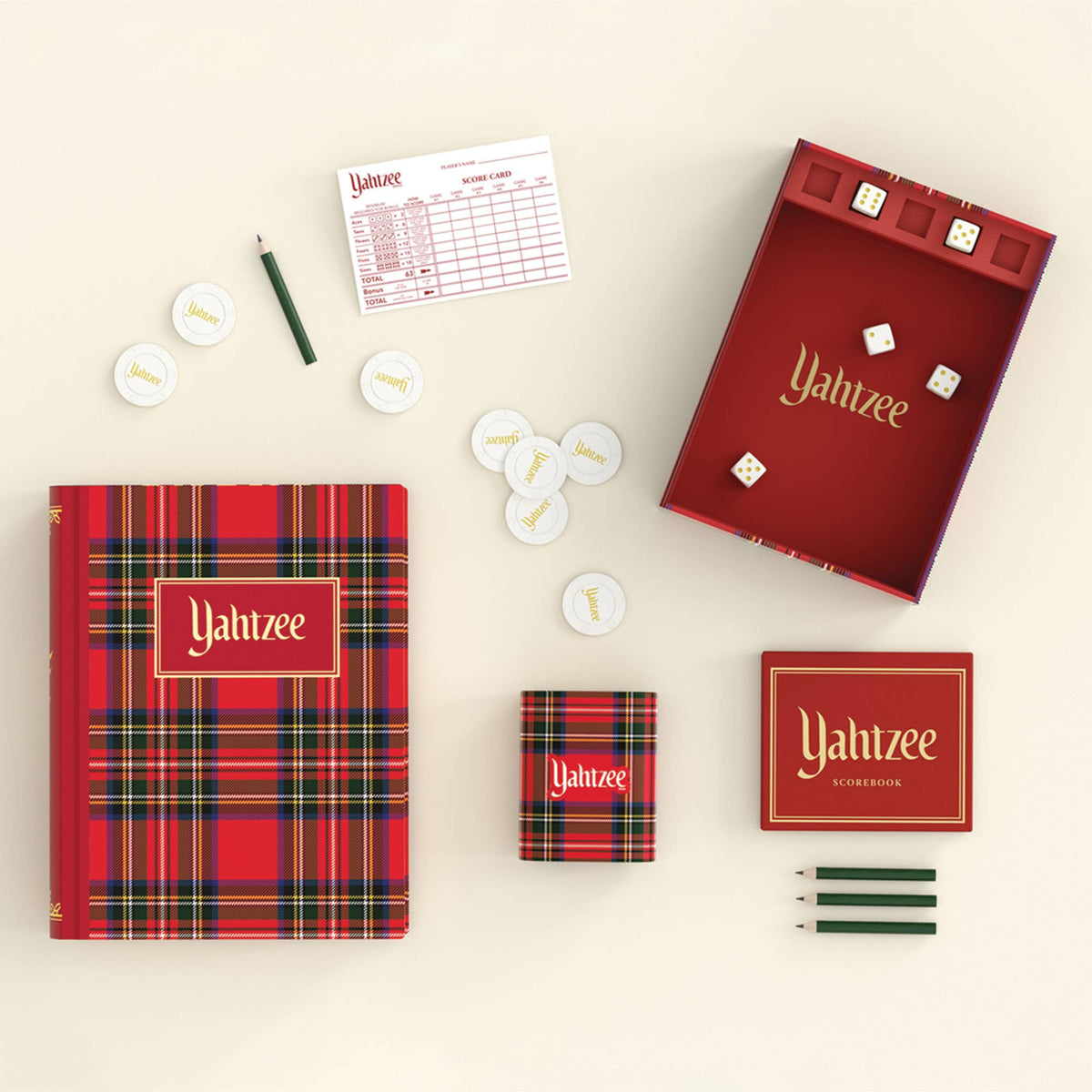 A flat-lay product shot of the Yahtzee game set on a warm cream background, including a red plaid game box, an open red plaid inner box with gold lettering, white scoring discs, a scoring sheet, three green pencils, and a small matching accessory box.