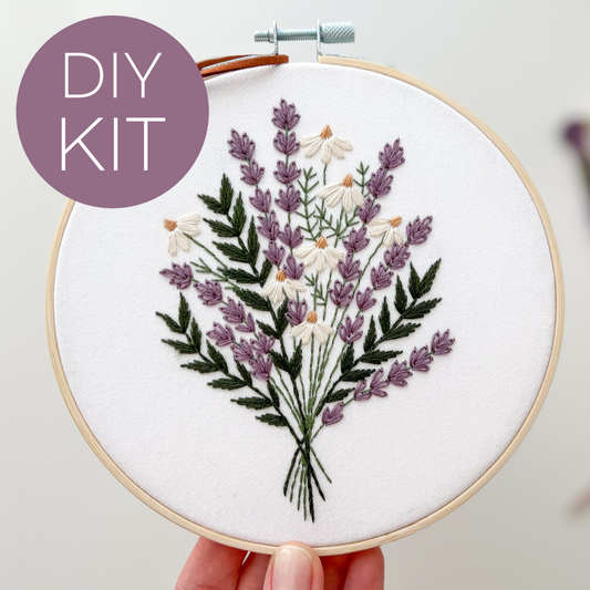 A hand holds a wooden embroidery hoop containing a floral embroidery design of lavender and white flowers with green stems. A purple circular badge in the top left reads 'DIY KIT'.