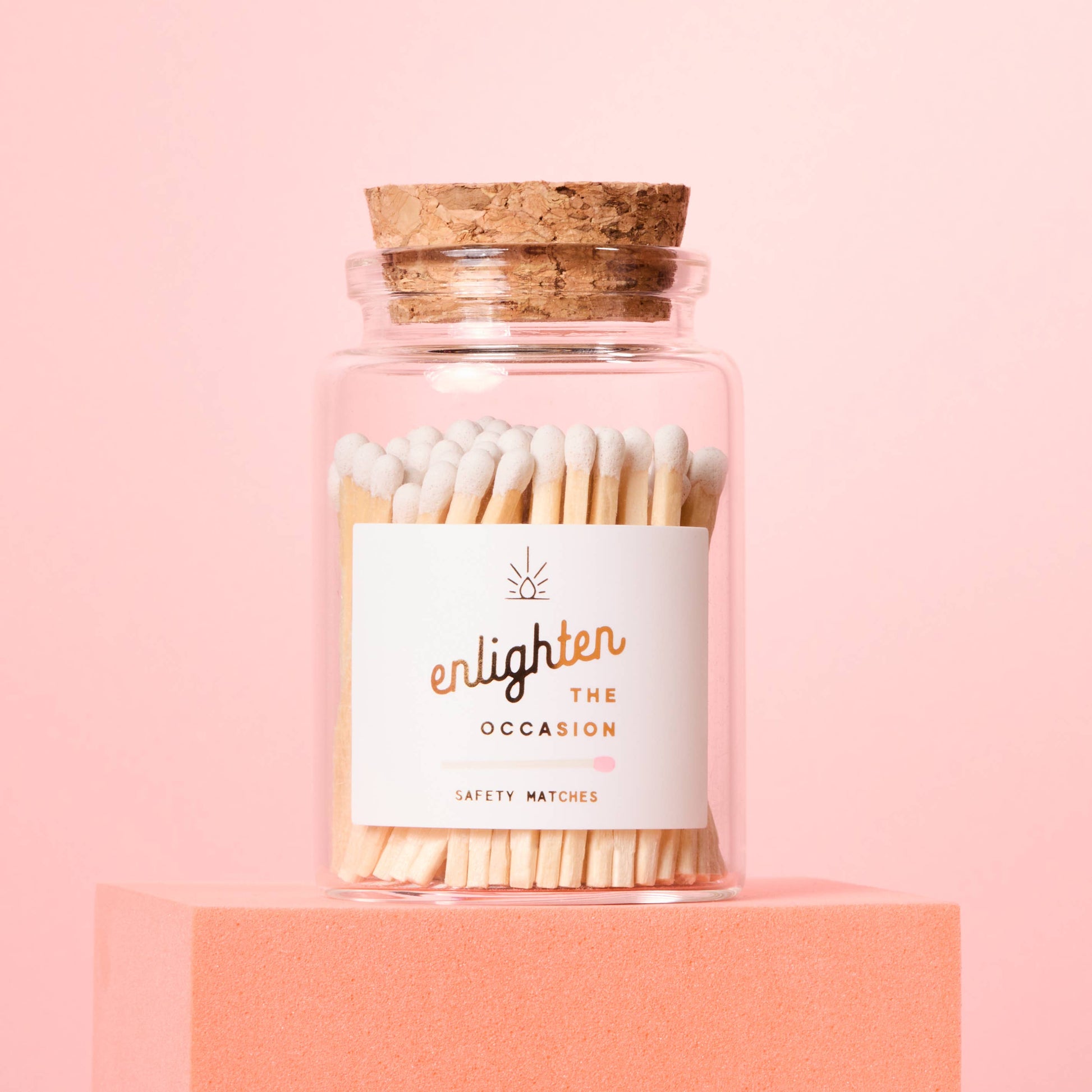 Jar of matches with a cork lid on a pink background, featuring the brand 'enlighten the occasion'.