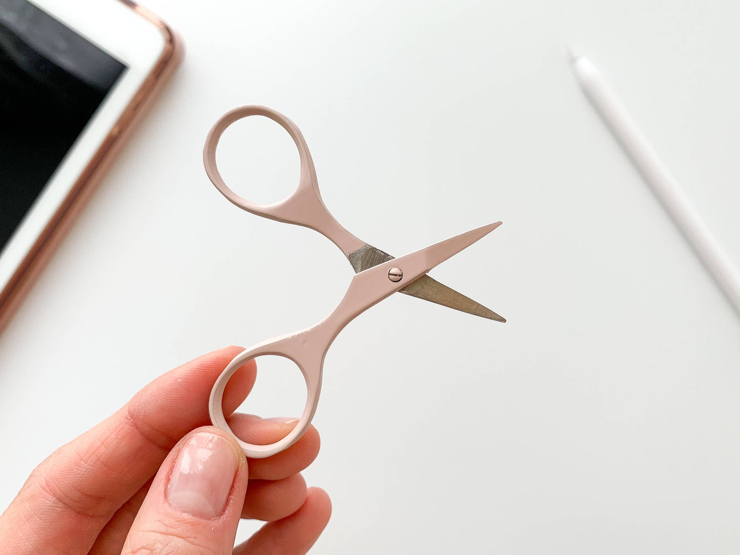 Small scissors held in a hand with a smartphone and pen in the background on a light surface