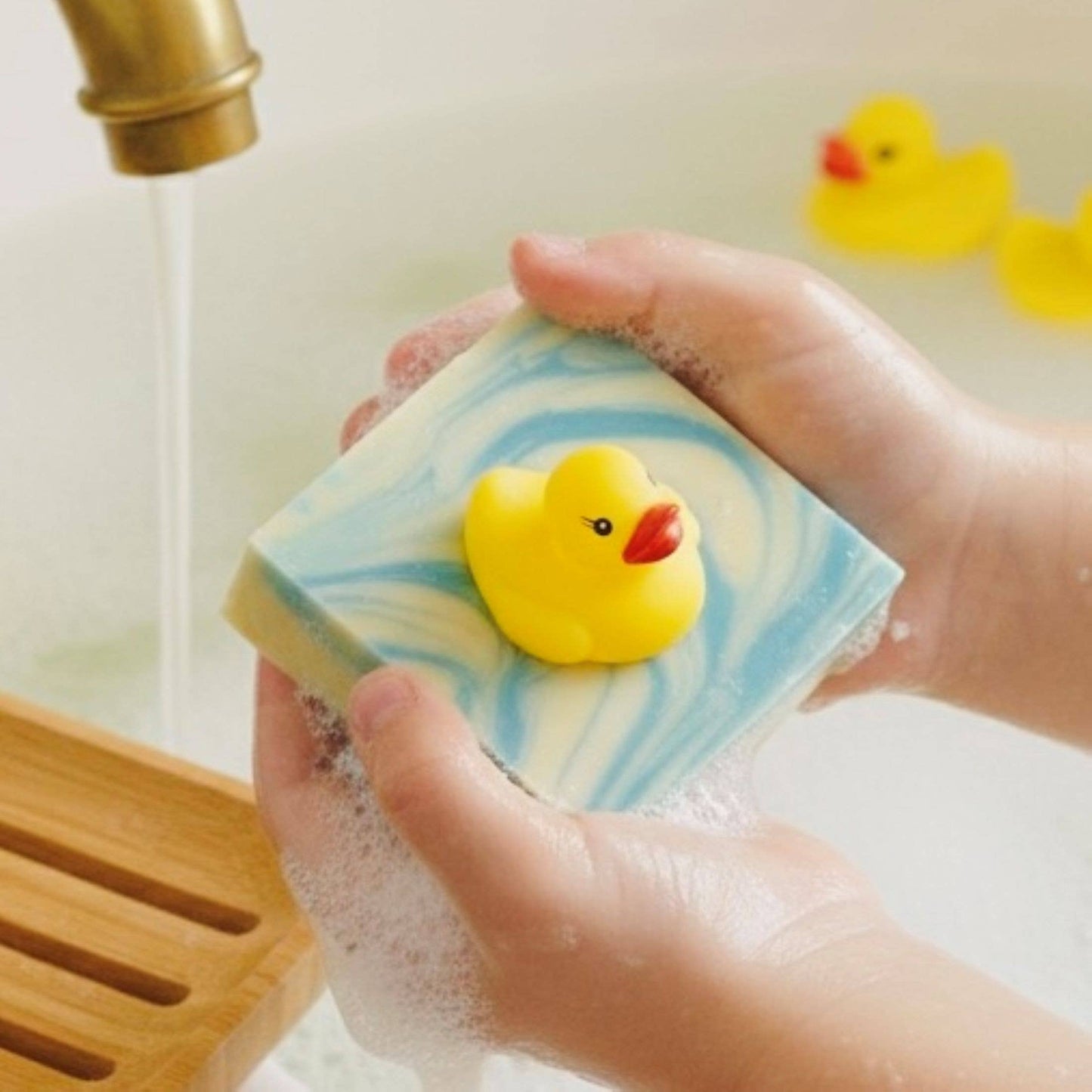 Hand holding a bar of soap with a yellow rubber duck on top, under running water.