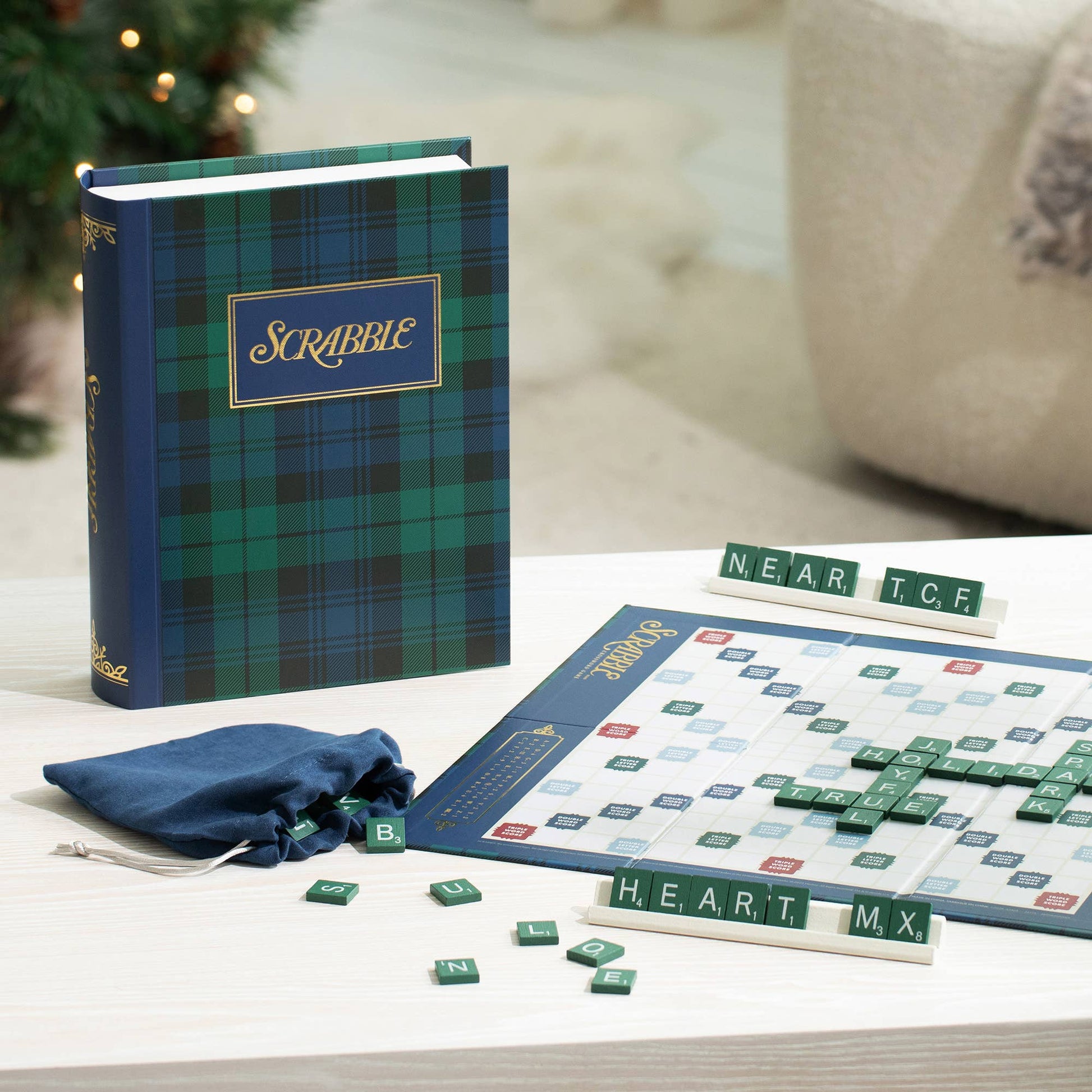 A personalized Scrabble word game set displayed on a light wood table, featuring a navy blue and green tartan patterned game book, a blue drawstring bag, a game board, and green letter tiles.