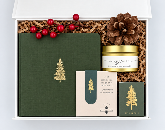 A curated gift box set on a white background, featuring a dark green journal with gold tree print, a pinecone, red berries, a jar labeled evergreen, a full-embossed magnetic bookmark, and a blue spruce card.
