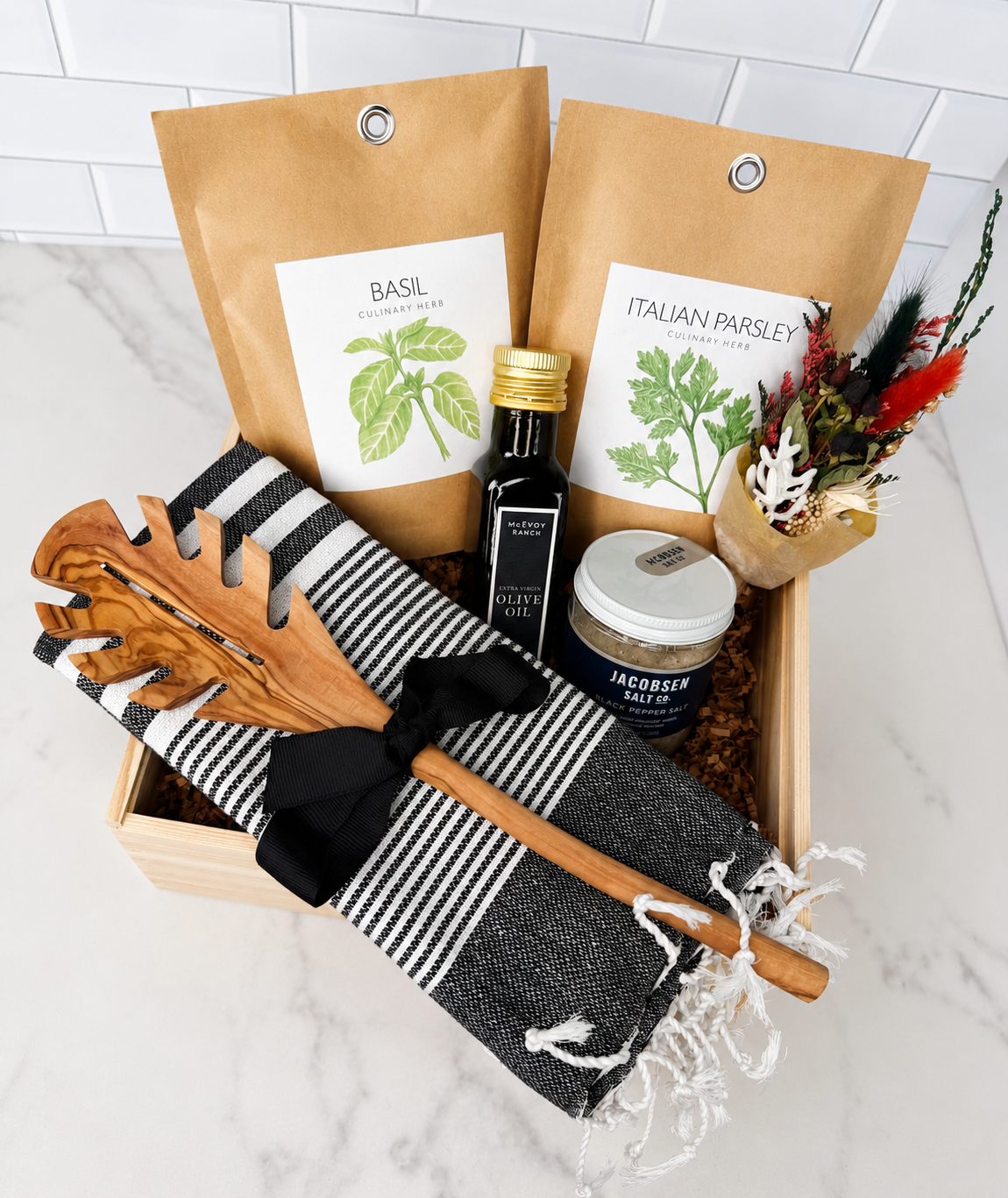 A curated gift basket set containing two bags of dried herbs (Basil and Italian Parsley), a bottle of olive oil, a jar of rosemary butter, a black and white striped kitchen towel, wooden utensils, and a small dried flower arrangement, all arranged in a wooden tray on a marble countertop.