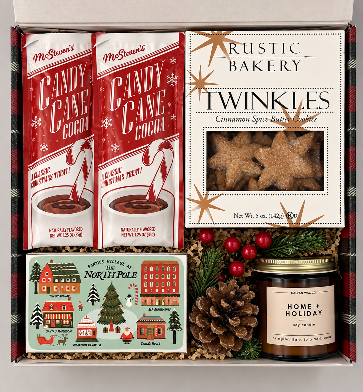 A curated holiday gift box containing multiple products: two packages of McSorran's Candy Cane Cocoa, a box of Rustic Bakery Twinkles, a Santa Village of the North Pole tin, and a Calm Down Wax Co. candle, arranged in a gift box with pinecones and red berries.