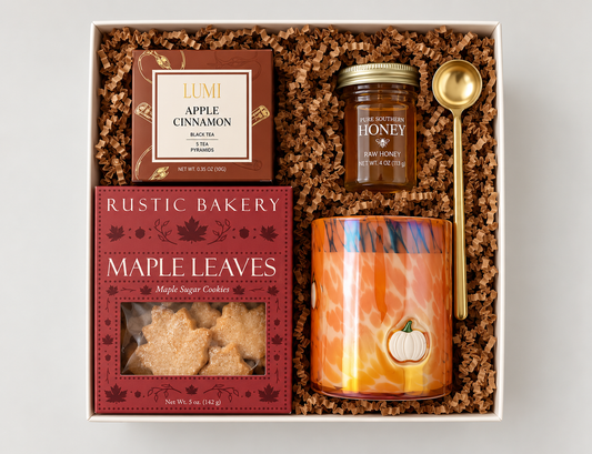 A curated gift box set containing a jar of Raw Honey from Four Sisters Farmhouse, a box of Lumi Apple Cinnamon Black Tea, a bag of Rustic Bakery Maple Leaves cookies, and an orange maple-themed scented candle, all arranged in brown shredded paper inside a white box.