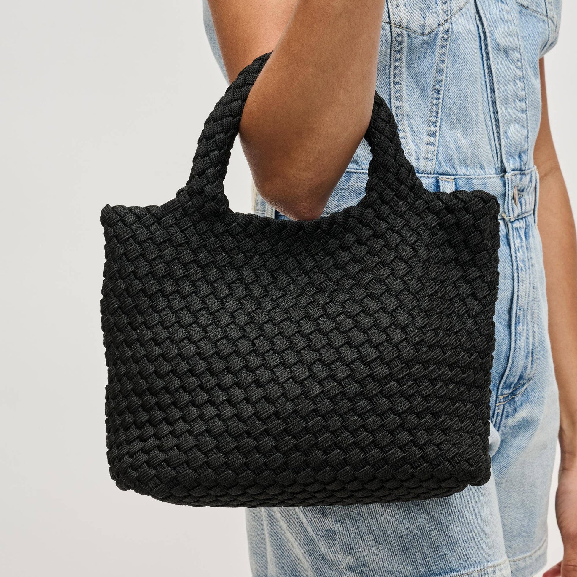 Black woven handbag held by a person wearing light blue jeans on a plain background