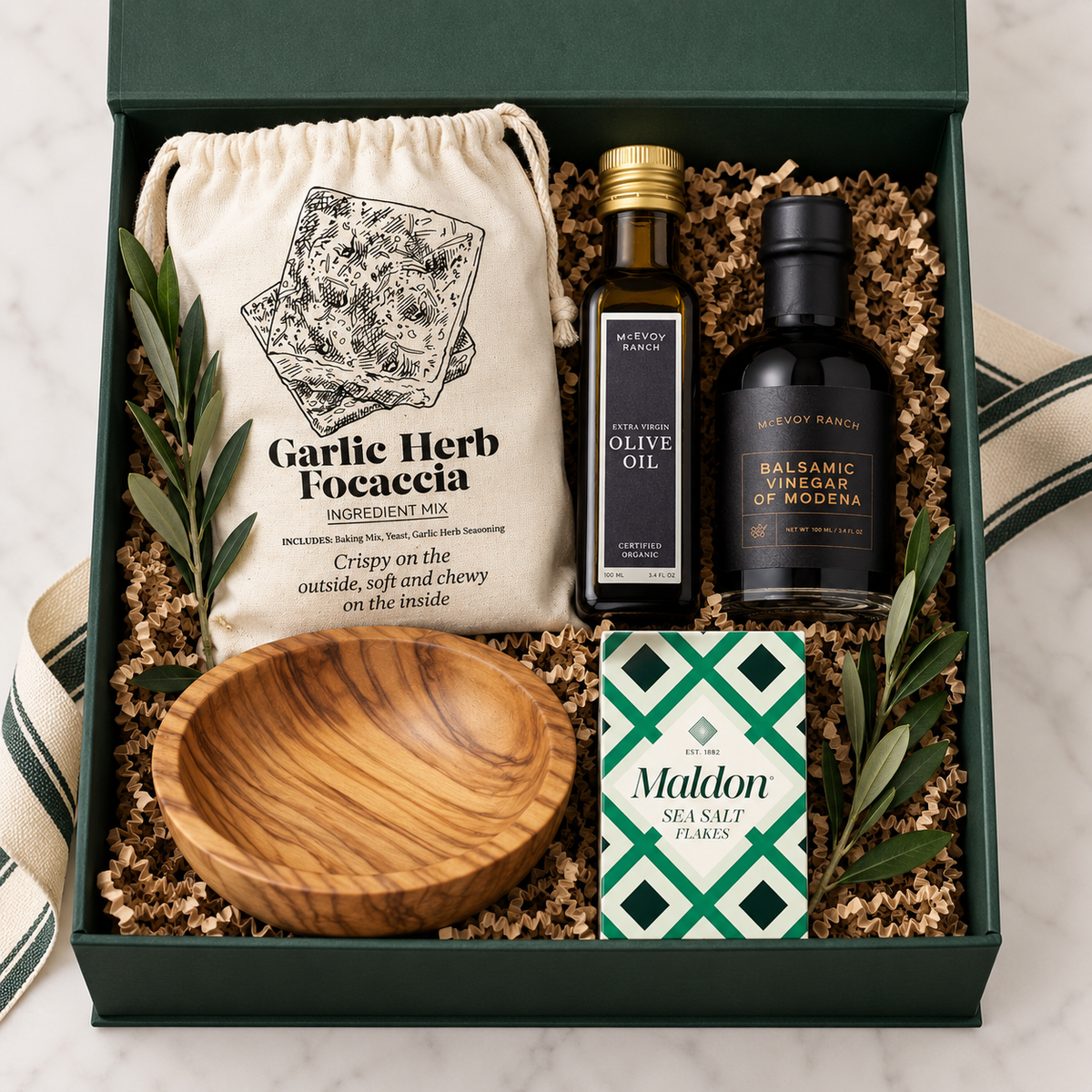 A gourmet gift box set containing a bag of Garlic Herb Focaccia mix, a bottle of Olive Oil, a bottle of Balsamic Vinegar of Modena, a box of Maldon Sea Salt, and a wooden bowl, all arranged in a dark green box with shredded paper filler.