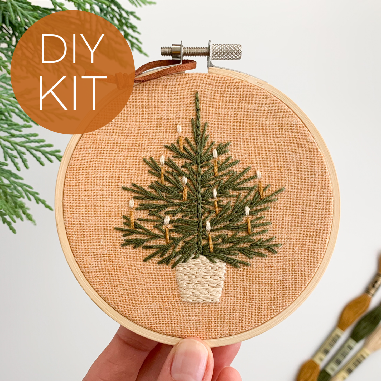 Embroidery hoop with a Christmas tree design held by a hand, surrounded by greenery and paintbrushes.