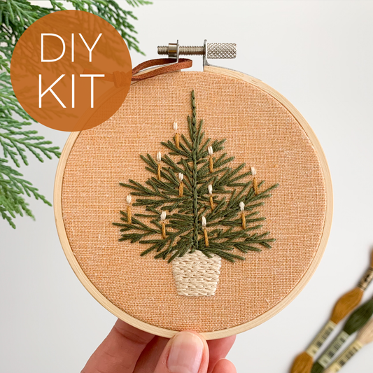Embroidery hoop with a Christmas tree design held by a hand, surrounded by greenery and paintbrushes.