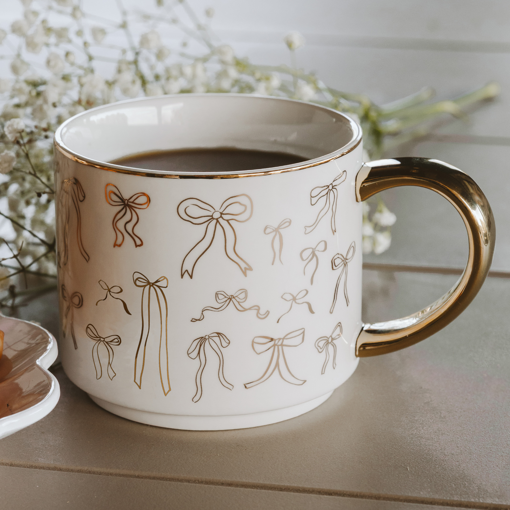 White mug with gold handle and bow pattern filled with coffee on a light surface.