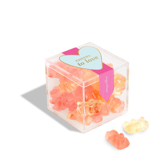 Clear cube box with pink and blue heart-shaped label containing small orange and yellow items on a white background