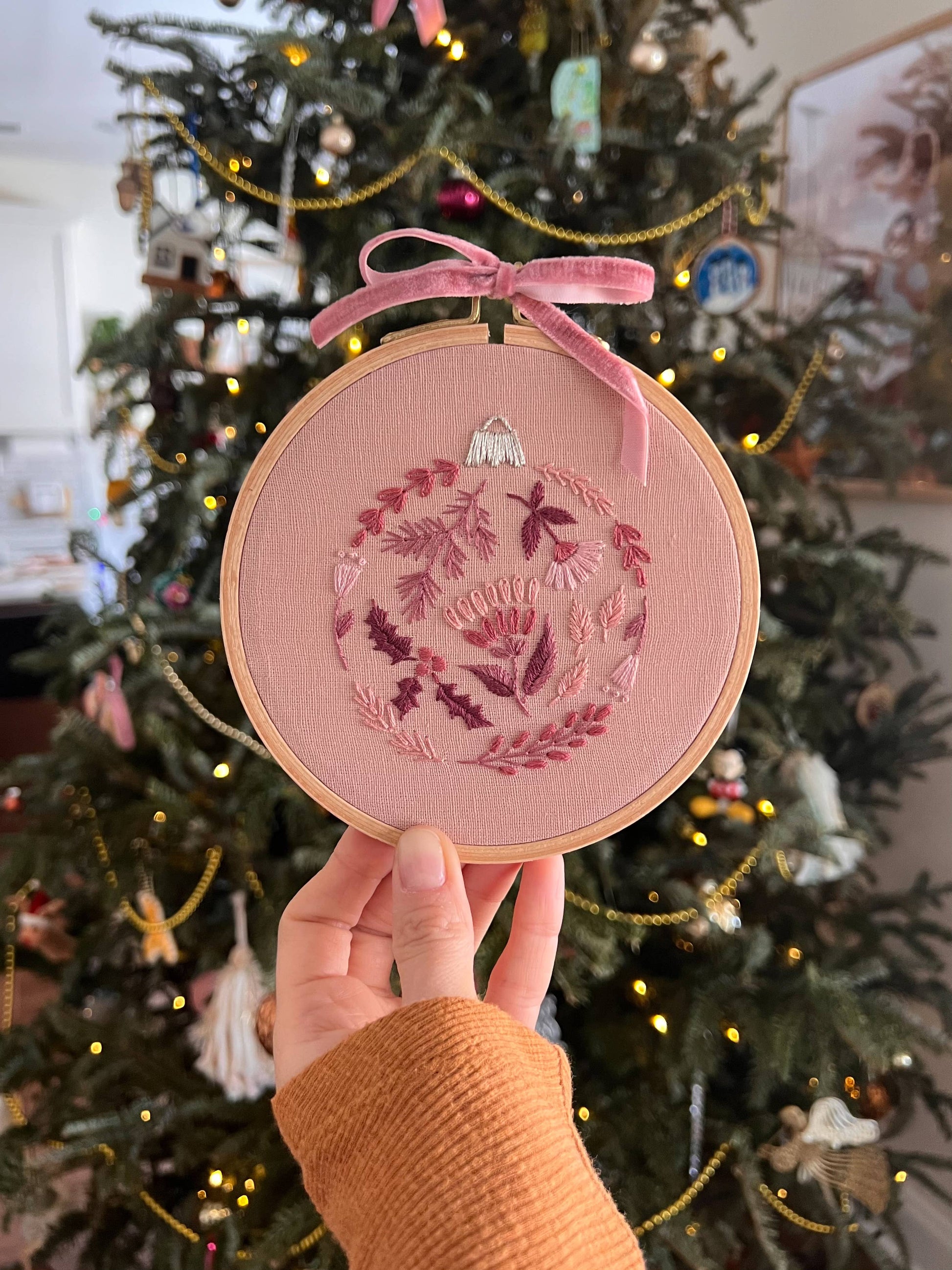 Embroidered pink ornament with floral design held in front of a decorated Christmas tree.