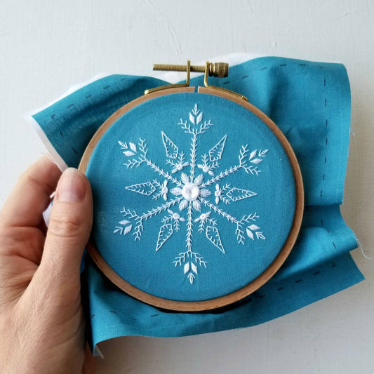 Embroidery hoop with a snowflake design on blue fabric held by a hand.