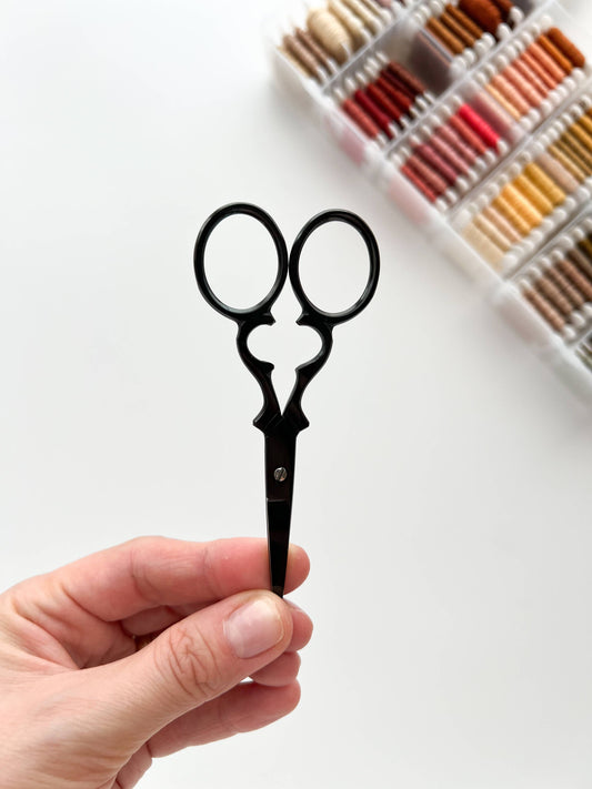 A hand holding a pair of small black scissors against a white background, with a blurred tray of colorful thread or cord spools in the upper right.