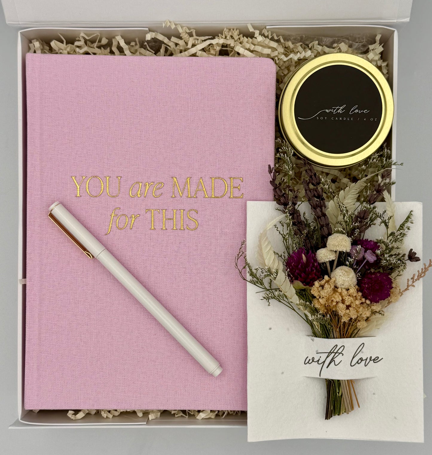 Pink notebook with a pen, small round item, and dried flowers in a box with 'You are Made for This' text.