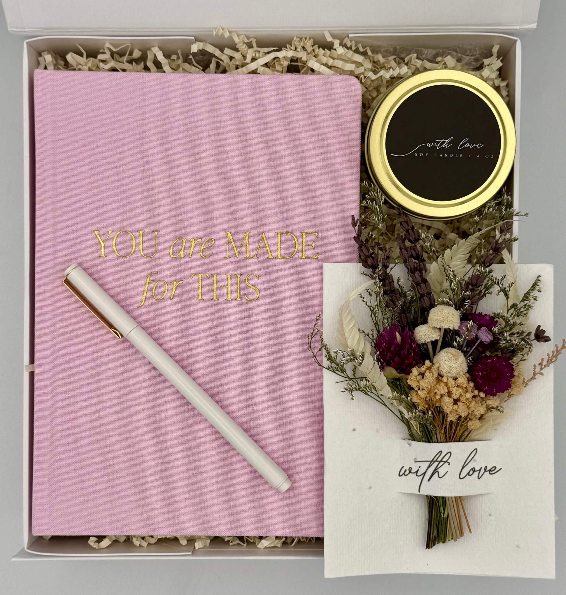 Pink notebook with a pen, small round item, and dried flowers in a box with 'You are Made for This' text.