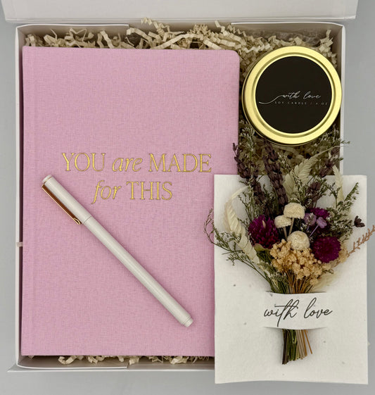 Pink notebook with a pen, small round item, and dried flowers in a box with 'You are Made for This' text.