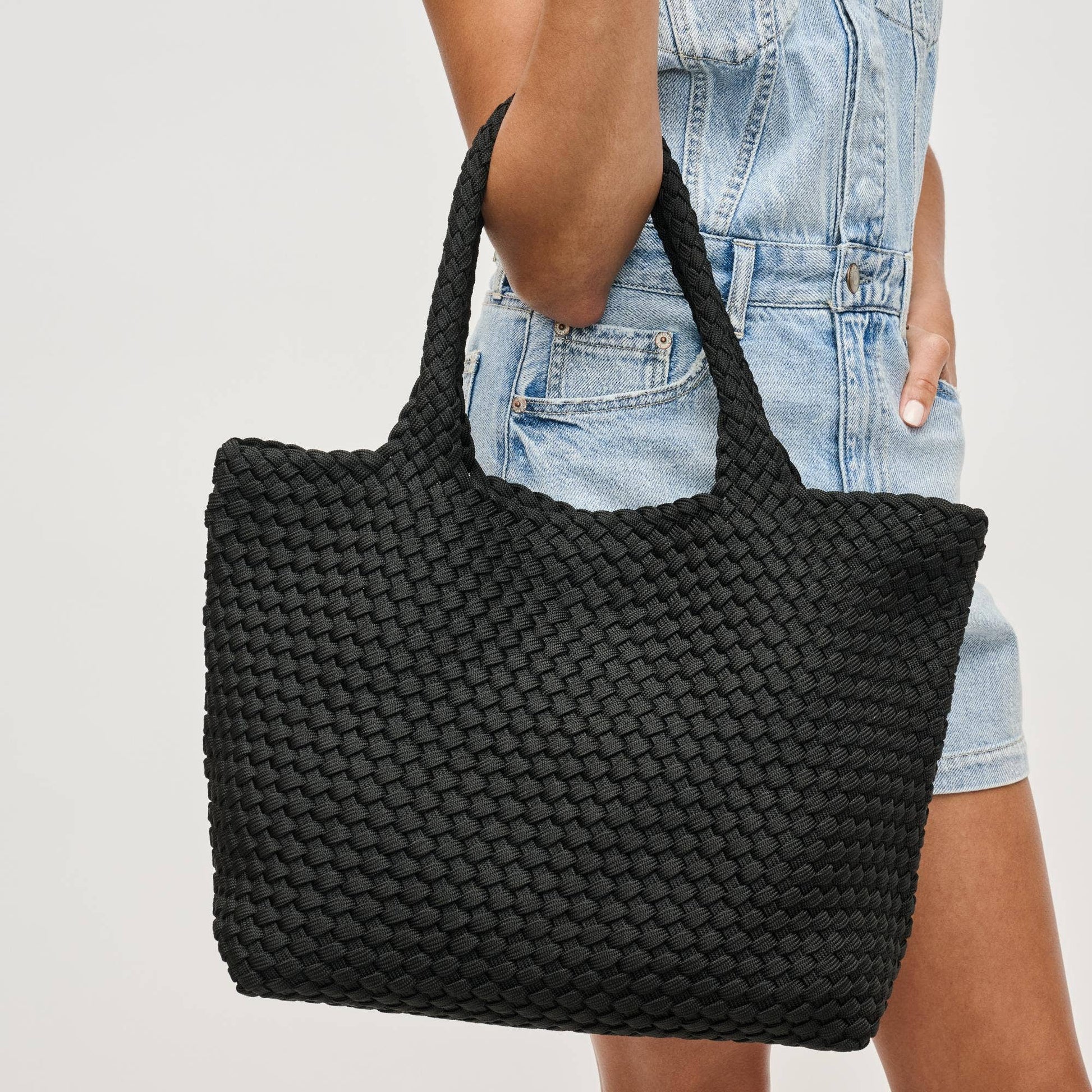 Black woven tote bag held by a person wearing a denim outfit on a light gray background